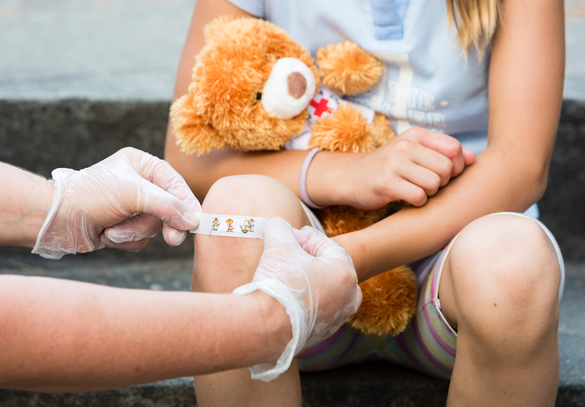 Kind hält Teddybär mit rotem Kreuz, während jemand mit Handschuhen ein Pflaster auf das Knie klebt, Erste Hilfe DRK