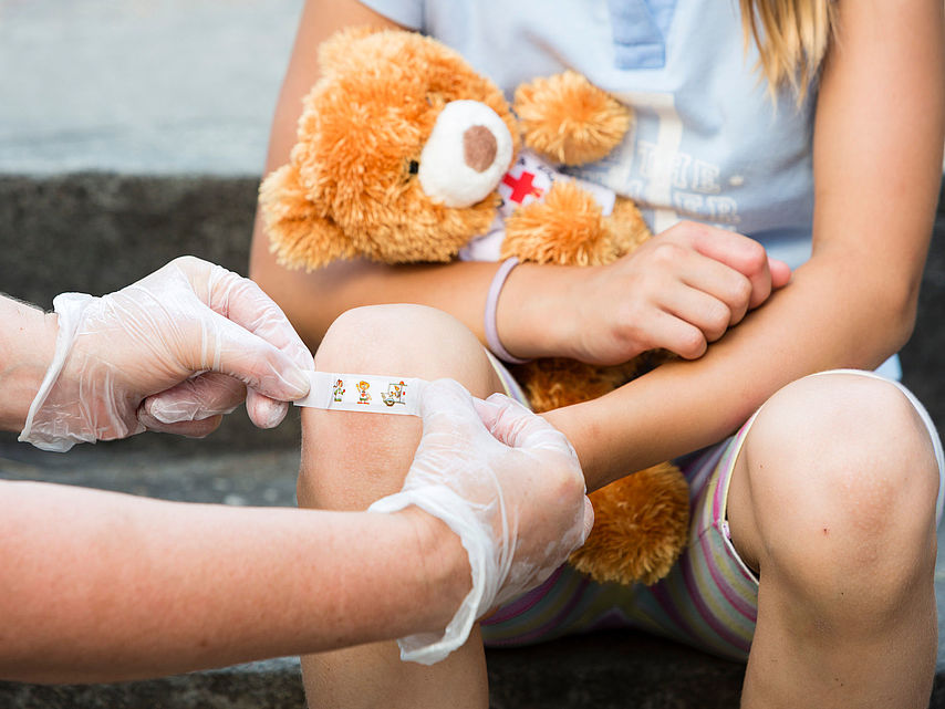 Kind hält Teddybär mit rotem Kreuz, während jemand mit Handschuhen ein Pflaster auf das Knie klebt, Erste Hilfe DRK