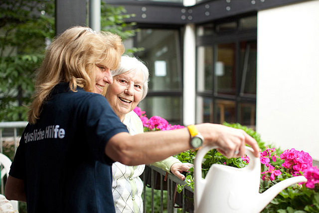 DRK-Mitarbeiterin der häuslichen Pflege hilft Seniorin pinke Blumen auf dem Balkon zu gießen