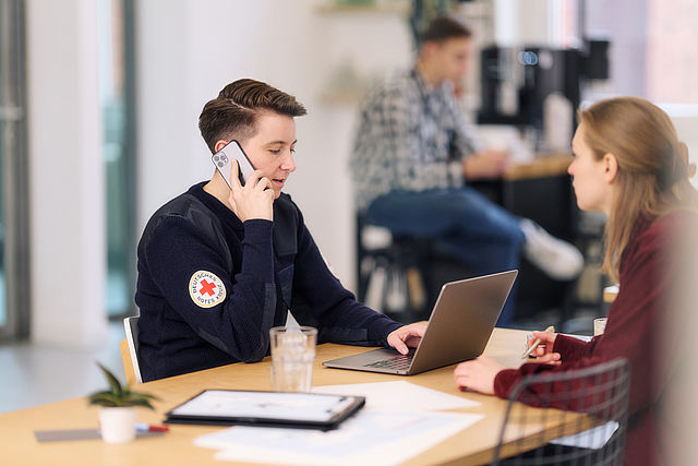 Symbolfoto, Mitarbeiterin des DRK arbeitet am Laptop und telefoniert mit einem Smartphone