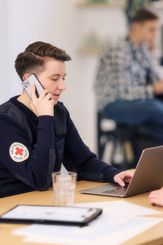 Symbolfoto, Mitarbeiterin des DRK arbeitet am Laptop und telefoniert mit einem Smartphone
