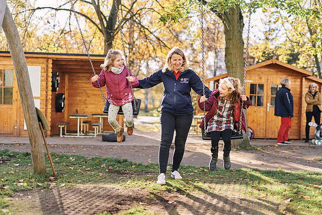 Zwei schaukelnde Kinder, in der Mitte steht eine Erzieherin