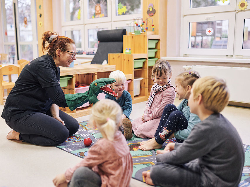 Erzieherin spielt mit einer Handpuppe (ein Dinosaurier) vor einer Gruppe Kinder