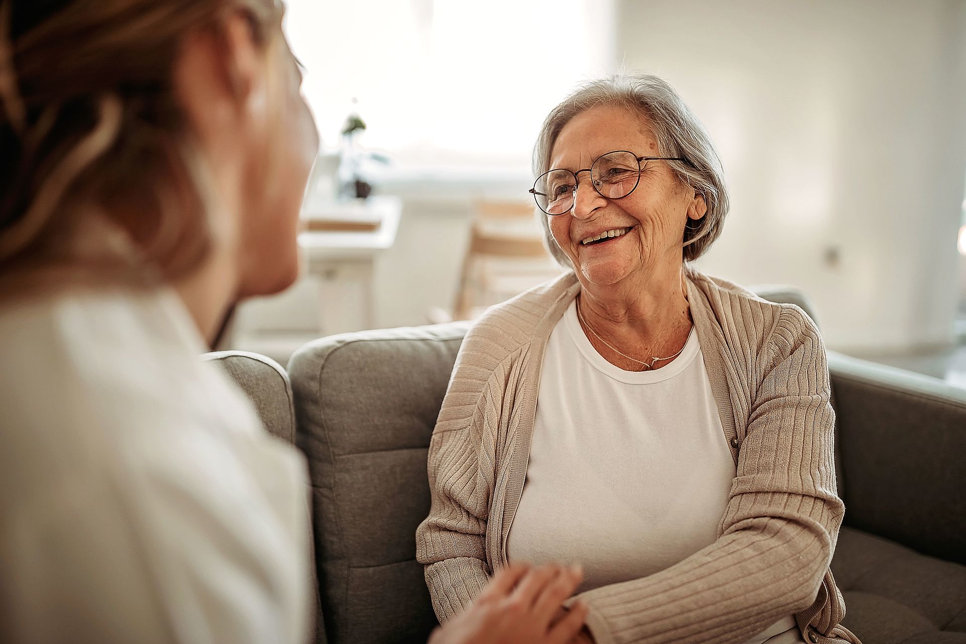 Eine lächelnde Seniorin sitzt auf einer Couch und ist im Gespräch mit einer jüngeren Erwachsenen.